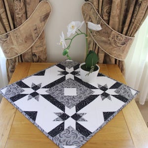 May include: A black and white quilt-style table runner with a starburst pattern. The runner is placed on a wooden table, with a white potted plant and curtains in the background. The runner measures approximately 24 inches square.