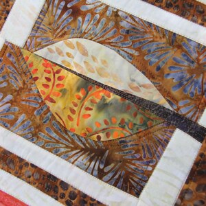 K&ouml;nnte beinhalten: Nahaufnahme eines Quilts mit einem Blattdesign. Der Quilt zeigt eine Kombination aus braunen, orangefarbenen und blauen Stoffen. Das Blatt besteht aus orangefarbenem und gelbem Stoff mit einem braunen Rand. Wei&szlig;e R&auml;nder umrahmen das Blatt und andere Abschnitte.