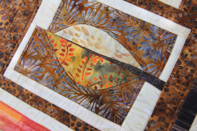 K&ouml;nnte beinhalten: Nahaufnahme eines Quilts mit einem Blattdesign. Der Quilt zeigt eine Kombination aus braunen, orangefarbenen und blauen Stoffen. Das Blatt besteht aus orangefarbenem und gelbem Stoff mit einem braunen Rand. Wei&szlig;e R&auml;nder umrahmen das Blatt und andere Abschnitte.