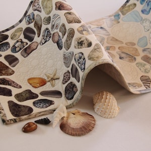 May include: A decorative fabric with a pebble and seashell design. The fabric features various colors of pebbles, seashells, and starfish. Several seashells are placed in front of the fabric.