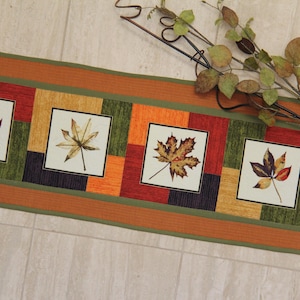 Puede incluir: Un camino de mesa rectangular de tela con un diseño de patchwork de cuadrados naranjas, verdes, amarillos y morados oscuros. El camino incluye paneles cuadrados blancos, cada uno con una ilustración detallada de una hoja de otoño.