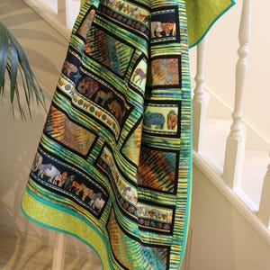 May include: A colorful quilt with an animal print design, featuring elephants, zebras, and other wildlife. The quilt has a green border and is draped over a wooden ladder. The color palette includes green, yellow, black, and orange.