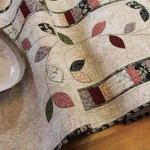 May include: A beige quilted table runner with a leaf design and a ceramic bowl. The runner features a floral and leaf pattern in shades of red, green, and black. The bowl is light beige with a speckled texture.