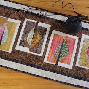 May include: A quilted table runner with four panels, each with a leaf design in pink, green, orange, and yellow. The runner has a brown and white border. A black teapot sits on the wooden surface.