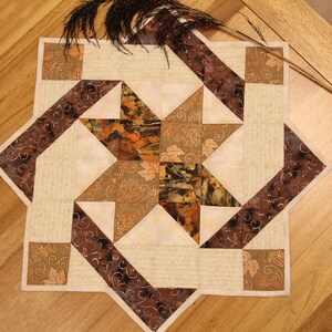 May include: A square fabric quilt topper featuring a central star pattern in shades of brown, tan, and cream. The design incorporates leaf motifs and swirling patterns, with dark brown borders and light cream background fabric sections.