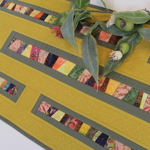 May include: Mustard yellow quilted table runner with geometric design. The runner features rectangular sections of colorful, patterned fabric. A sprig of greenery with leaves and small green fruit rests on the runner.