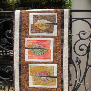 K&ouml;nnte beinhalten: Ein handgefertigter Wandbehang mit drei rechteckigen Tafeln mit abstrakten Blattmotiven. Der Quilt hat ein braunes und cremefarbenes Farbschema mit einem gemusterten Rand und einer cremefarbenen R&uuml;ckseite. Die Blattmotive enthalten Farbt&ouml;ne von Orange, Gr&uuml;n und Gelb.