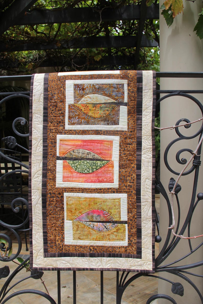 K&ouml;nnte beinhalten: Ein handgefertigter Wandbehang mit drei rechteckigen Tafeln mit abstrakten Blattmotiven. Der Quilt hat ein braunes und cremefarbenes Farbschema mit einem gemusterten Rand und einer cremefarbenen R&uuml;ckseite. Die Blattmotive enthalten Farbt&ouml;ne von Orange, Gr&uuml;n und Gelb.