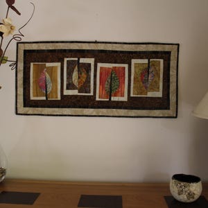 K&ouml;nnte beinhalten: Ein rechteckiges Wandbehang mit einem braunen und beigen Rand zeigt vier rechteckige Tafeln mit blattartigen Designs in Rot-, Gelb- und Gr&uuml;nt&ouml;nen. Eine klare Glasvase mit dekorativen Zweigen und eine Lampe mit gemustertem Sockel sind ebenfalls zu sehen.