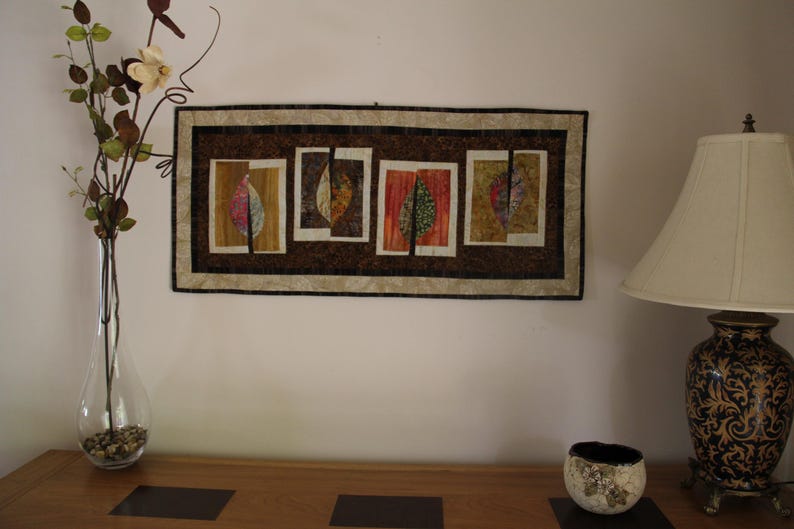 K&ouml;nnte beinhalten: Ein rechteckiges Wandbehang mit einem braunen und beigen Rand zeigt vier rechteckige Tafeln mit blattartigen Designs in Rot-, Gelb- und Gr&uuml;nt&ouml;nen. Eine klare Glasvase mit dekorativen Zweigen und eine Lampe mit gemustertem Sockel sind ebenfalls zu sehen.