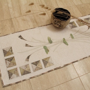 May include: A rectangular quilted table runner featuring a cream-colored fabric base with a central botanical design of green leaves and thin stems. The border includes a series of small square patchwork blocks in shades of brown and beige.