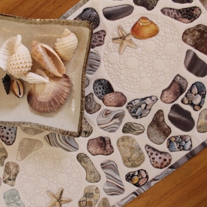 May include: A square ceramic dish filled with seashells rests on a beach-themed table runner. The runner displays a pattern of pebbles, starfish, and shells in neutral tones. The dish and runner create a coastal aesthetic.