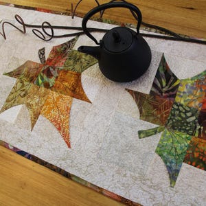 May include: A decorative table runner with large, colourful leaf designs in shades of green, orange, and brown. A small black teapot sits on the runner. The runner is approximately 30 cm wide.