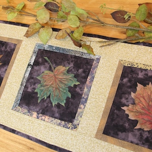 May include: A rectangular table runner with three square panels featuring colourful leaf designs. The leaves are in shades of red, orange, and green against a dark purple background. The runner has a beige border and a decorative leaf branch.