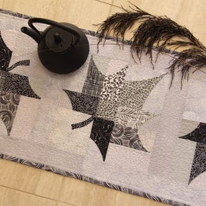 May include: A gray and white quilted table runner featuring three stylized maple leaf designs in black and white patterns. A black teapot sits on the runner, and a sprig of dried ornamental grass is placed on the right side.