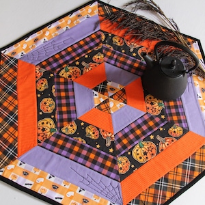 May include: A hexagonal Halloween-themed table runner with a black border. The runner features orange, purple, and black plaid and pumpkin patterns. A small black teapot sits on the runner, with dried decorative stems.