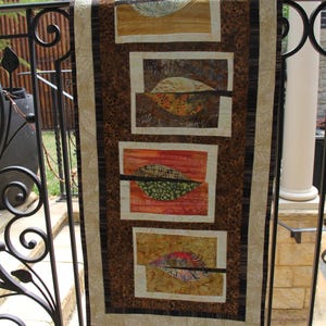 K&ouml;nnte beinhalten: Ein dekorativer Wandbehang mit einem von der Natur inspirierten Design. Der Stoff hat vier rechteckige Paneele mit blattartigen Formen in Orange-, Gr&uuml;n- und Gelbt&ouml;nen. Der Hintergrund hat ein braunes Leopardenmuster und die R&auml;nder sind mit einem beigen Rand versehen.