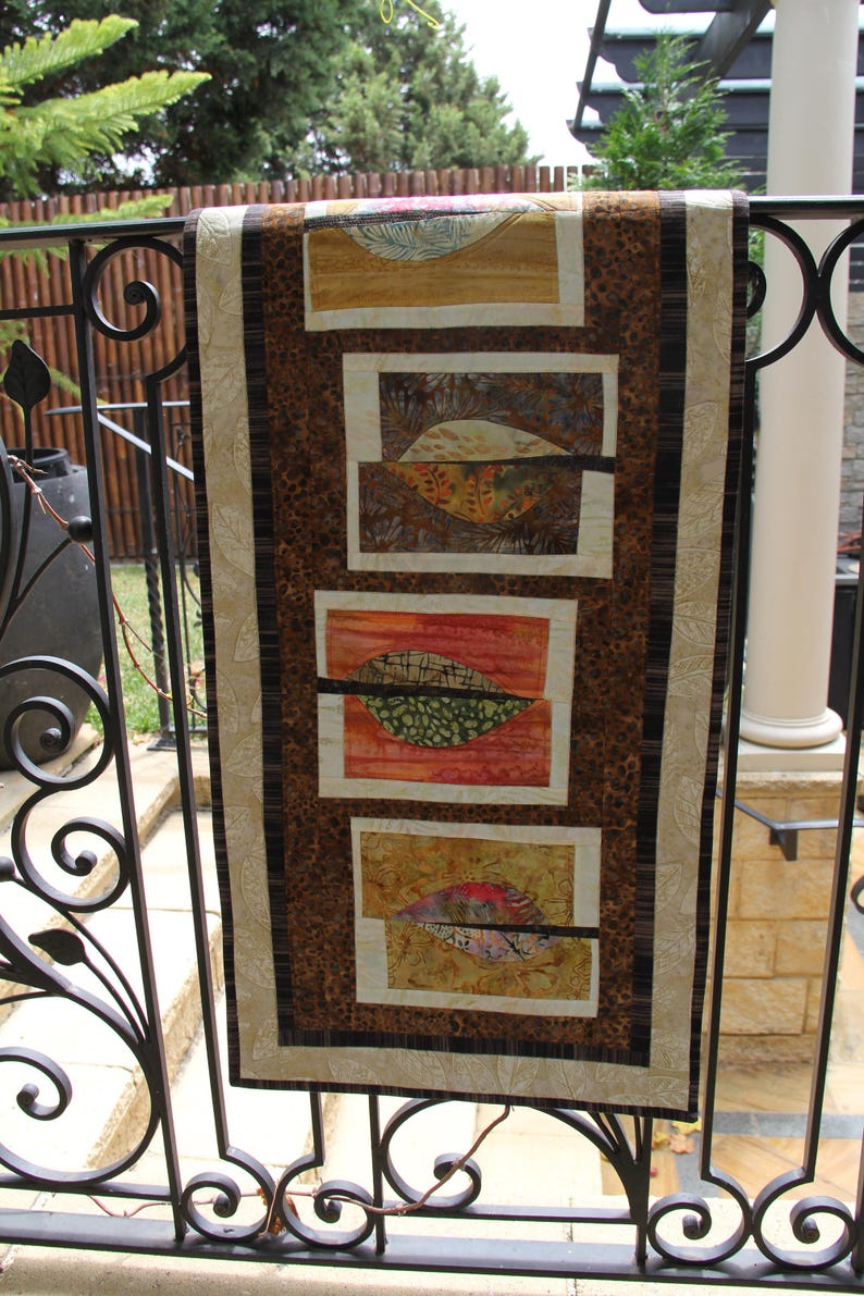 K&ouml;nnte beinhalten: Ein dekorativer Wandbehang mit einem von der Natur inspirierten Design. Der Stoff hat vier rechteckige Paneele mit blattartigen Formen in Orange-, Gr&uuml;n- und Gelbt&ouml;nen. Der Hintergrund hat ein braunes Leopardenmuster und die R&auml;nder sind mit einem beigen Rand versehen.