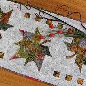 May include: A quilted table runner with three large, colourful leaf shapes in shades of green, orange, and brown. The leaves are set against a light grey background with small square accents. A decorative twig with red berries and a brown metal swirl accent the runner.
