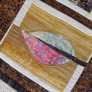 K&ouml;nnte beinhalten: Nahaufnahme eines gesteppten Textils mit einem blattartigen Design. Der Quilt hat eine zentrale Form mit rosa, blauen und gr&uuml;nen Mustern, vor einem beigen Hintergrund. Der Quilt hat einen braunen und wei&szlig;en Rand.