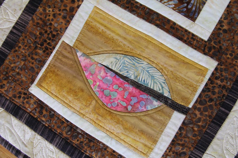 K&ouml;nnte beinhalten: Nahaufnahme eines gesteppten Textils mit einem blattartigen Design. Der Quilt hat eine zentrale Form mit rosa, blauen und gr&uuml;nen Mustern, vor einem beigen Hintergrund. Der Quilt hat einen braunen und wei&szlig;en Rand.