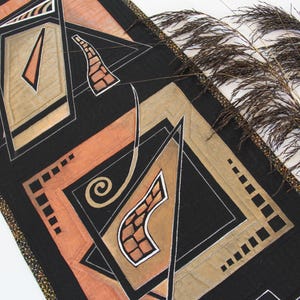 May include: A black and tan geometric design table runner with a gold trim. The runner features abstract shapes in shades of tan, peach, and black. Decorative dried plants are in the background.