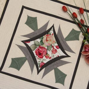 Op de afbeelding: Een vierkant stoffen quiltblok met een centrale ruitvorm en een bloemenpatroon van rozen in rood, roze en groen. Het blok is omlijst door een zwarte rand en geaccentueerd met grijze en groene bladvormige stoffen applicaties op een crèmekleurige achtergrond.