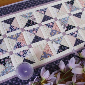 May include: A rectangular quilted table runner featuring a patchwork design of triangular fabric pieces in shades of purple, pink, and white with floral patterns, bordered by a dark purple fabric with a small white floral print.