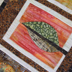 K&ouml;nnte beinhalten: Nahaufnahme eines Quilts mit einem Blattdesign. Der Quilt hat ein zentrales Quadrat mit einer Blattform in Gr&uuml;n und Braun, vor einem roten und orangen Hintergrund. Der Quilt hat einen wei&szlig;en Rand und einen braunen gemusterten Rand.