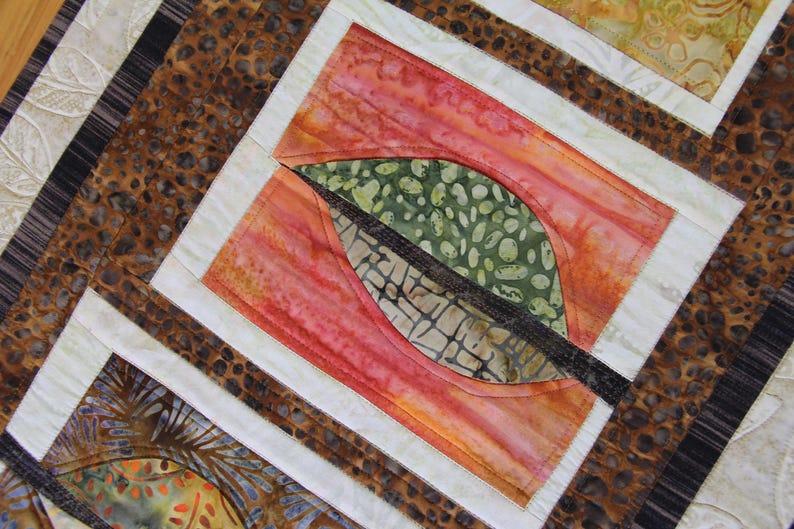 K&ouml;nnte beinhalten: Nahaufnahme eines Quilts mit einem Blattdesign. Der Quilt hat ein zentrales Quadrat mit einer Blattform in Gr&uuml;n und Braun, vor einem roten und orangen Hintergrund. Der Quilt hat einen wei&szlig;en Rand und einen braunen gemusterten Rand.