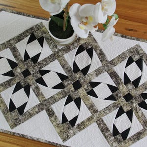 May include: A quilted table runner with a geometric pattern in black, white, and grey. The design features repeating squares with diamond and triangle shapes. A small white flower pot with white flowers is in the background.