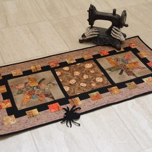 Può includere: Un runner da tavola rettangolare in tessuto con un design patchwork a tema autunnale, che include foglie d'acero e piccole zucche nei toni dell'arancione, marrone e nero, bordato da una sottile cornice nera e un bordo marrone screziato.
