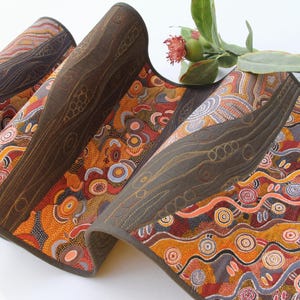 May include: A long, rectangular fabric table runner featuring a vibrant dot-art pattern in shades of orange, red, yellow, and brown, interspersed with solid dark brown panels that have intricate, quilted, swirling line designs throughout.