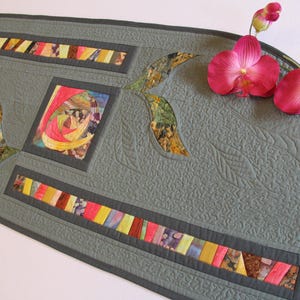 May include: A grey quilted table runner with colourful patchwork accents and a dark grey border. The runner features a central square and two rectangular sections with vibrant fabric strips. Pink orchids add a touch of colour.