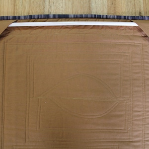K&ouml;nnte beinhalten: Nahaufnahme der R&uuml;ckseite eines braunen, gesteppten Wandbehangs. Der Quilt zeigt ein Blattmuster und geometrische Muster. Die Kanten sind mit einem blau-grau gestreiften Stoff versehen, und eine wei&szlig;e Stange ist zum Aufh&auml;ngen angebracht.