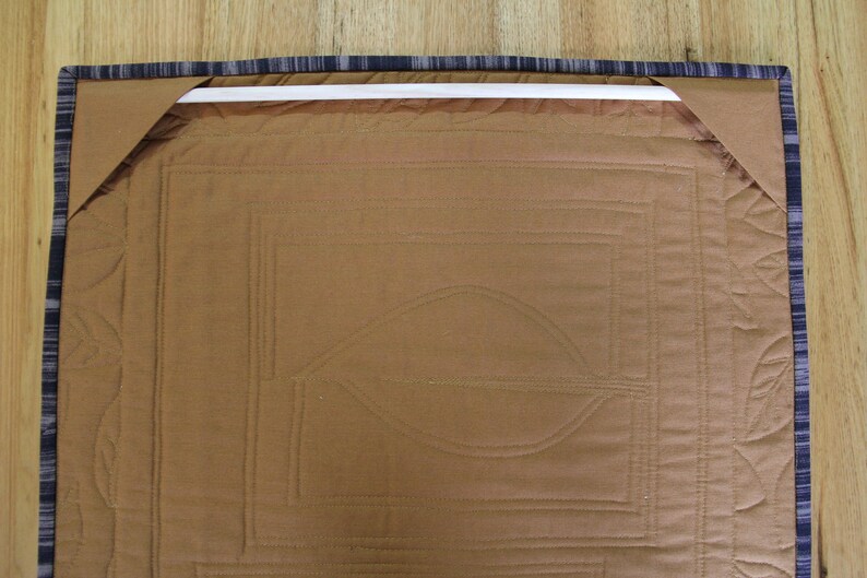 K&ouml;nnte beinhalten: Nahaufnahme der R&uuml;ckseite eines braunen, gesteppten Wandbehangs. Der Quilt zeigt ein Blattmuster und geometrische Muster. Die Kanten sind mit einem blau-grau gestreiften Stoff versehen, und eine wei&szlig;e Stange ist zum Aufh&auml;ngen angebracht.