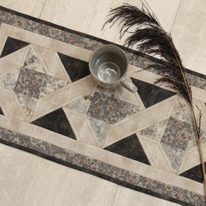 May include: A rectangular quilted table runner featuring a geometric star pattern in shades of beige, black, and grey with intricate floral details. A grey ceramic mug and a long, dark, feathery decorative branch are placed on the runner.
