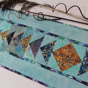 Op de afbeelding: Een turquoise gewatteerde tafelloper met een centraal ontwerp van kleurrijke geometrische vormen. De vormen omvatten driehoeken en vierkanten in tinten blauw, oranje en zwart. De loper is ongeveer 91 cm lang.