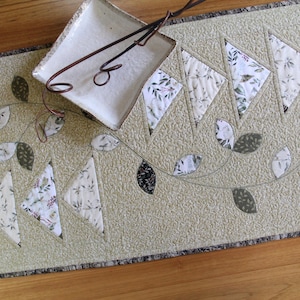 Könnte beinhalten: Beige gesteppter Tischläufer mit floralem Blattdesign. Der Läufer hat dreieckige Stoffformen mit floralem Muster und applizierten Blättern in Grün und Schwarz. Ein weißer quadratischer Teller und Metallzangen liegen obenauf.