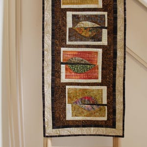 K&ouml;nnte beinhalten: Ein gestepptes Wandbehang mit einem Blattdesign. Das Kunstwerk zeigt rechteckige Paneele mit blattartigen Formen in Braun-, Rot- und Goldt&ouml;nen. Die R&auml;nder sind mit gemustertem Stoff und einem cremefarbenen Blattmuster eingefasst.