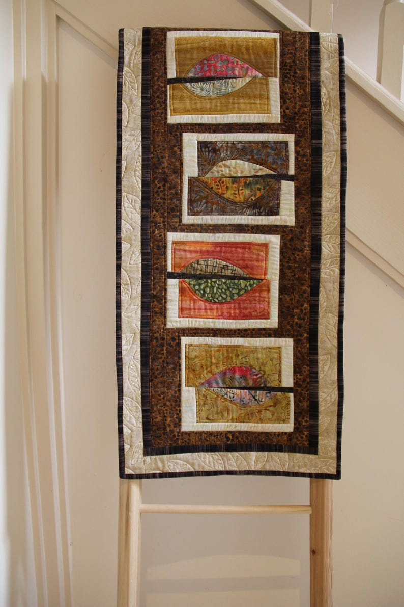 K&ouml;nnte beinhalten: Ein gestepptes Wandbehang mit einem Blattdesign. Das Kunstwerk zeigt rechteckige Paneele mit blattartigen Formen in Braun-, Rot- und Goldt&ouml;nen. Die R&auml;nder sind mit gemustertem Stoff und einem cremefarbenen Blattmuster eingefasst.