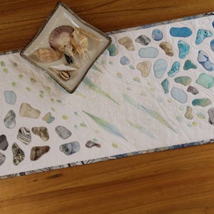 Könnte beinhalten: Ein rechteckiger, weißer Tischläufer mit maritimem Design. Er zeigt verschiedene Formen, die an Meeresglas in Blau-, Grün- und Beigetönen erinnern. Auf dem Läufer liegt eine quadratische Schale mit Muscheln. Der Läufer liegt auf einer Holzoberfläche.