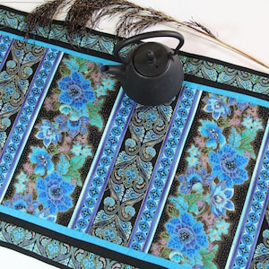 May include: A patterned table runner with a floral design in blue, teal, and black. The runner has a repeating pattern of flowers and geometric shapes. A black teapot sits on the runner.