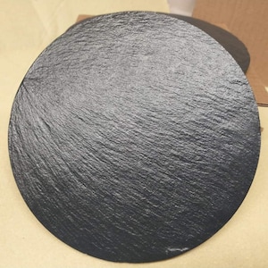 May include: Two round, dark grey slate serving platters with a textured surface. The platters are circular and appear to be made of natural stone. The slate has a rough, uneven texture, and the colour is a deep charcoal grey.