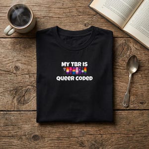 May include: Black t-shirt with the text "MY TBR IS queer coded" in white. The text is above a row of colorful book spines. A cup of coffee, a spoon, and an open book are on a wooden surface.