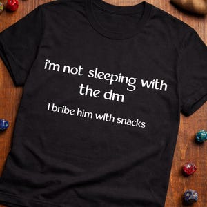 Op de afbeelding: Een witte keramische mok met een zwart handvat en de tekst "But First, Coffee" in een vetgedrukte, zwarte letter. De mok is gevuld met een donkere vloeistof, waarschijnlijk koffie, en staat op een lichtgekleurd oppervlak.