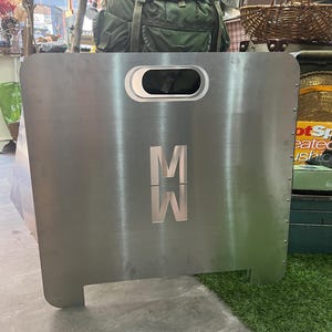 May include: A silver metal fire pit with a cutout in the shape of a circle. The fire pit has a metal grate and a metal stand. The fire pit has the letters "M" and "W" on the front.