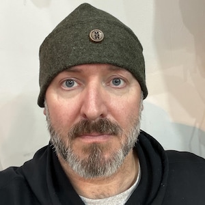 May include: A man wearing a dark green wool hat with a wooden button featuring the letters "M" and "W". He is wearing a black hooded sweatshirt.