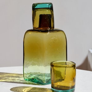 May include: A set of two glass containers. The larger, rectangular bottle has a teal top and base, with a yellow-green body. A smaller, cylindrical glass sits beside it, also with teal accents. The glasses cast shadows.