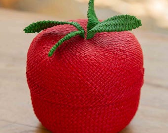 May include: A red, tomato-shaped decorative box with green leaves. The box has a woven texture and sits on a wooden surface. The leaves are a vibrant green, contrasting with the red body of the box.
